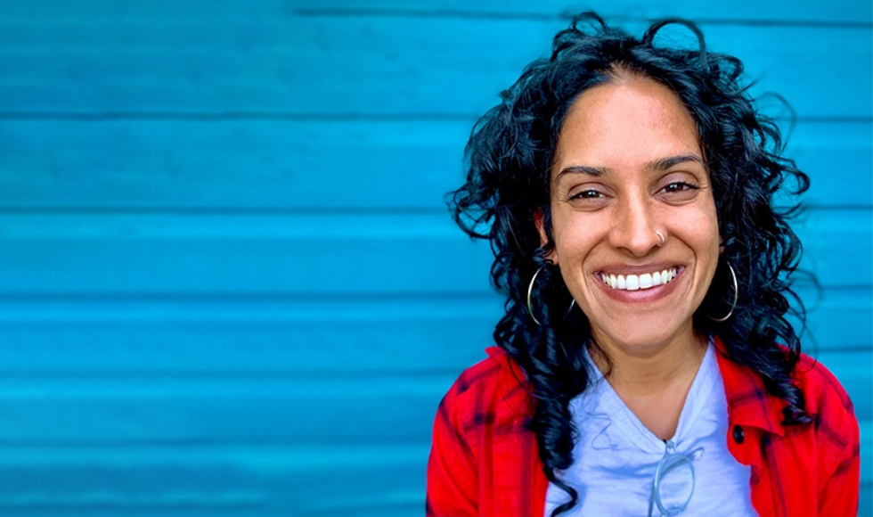 A close up shot of Anjali Menezes smiling at the camera. She is wearing a blue t-shirt, read jacket and is standing against a bright blue background.