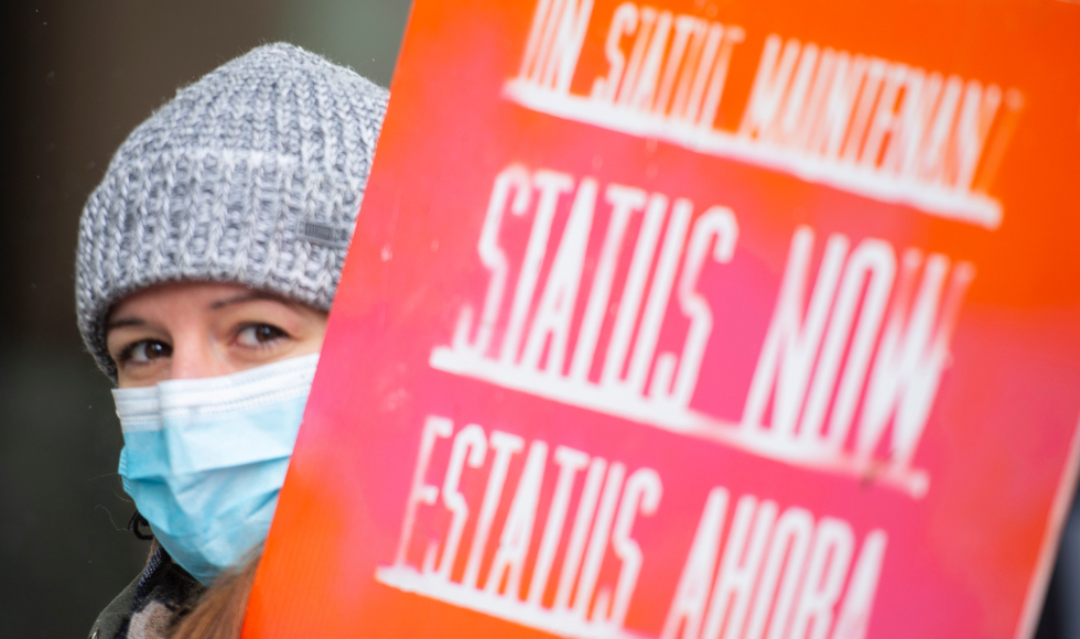 A person peeks out from behind a protest sign in the foreground, calling for status for undocumented migrants