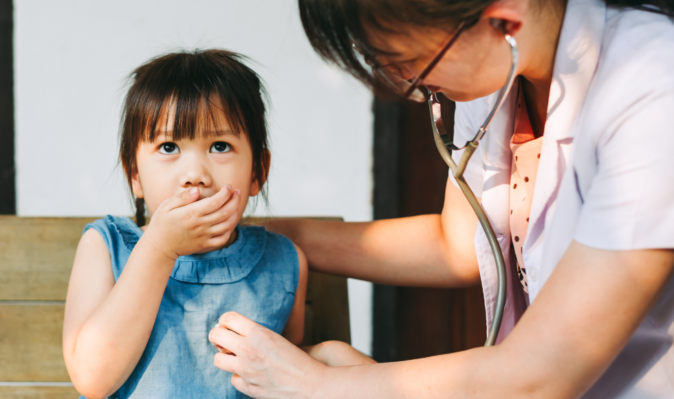 A child covering their mouth while a person in a lab coat listens to their chest with a stethescope.