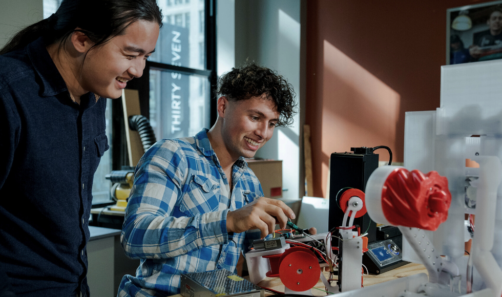 Cheng smiles as Owais works a machine on a table in front of them.