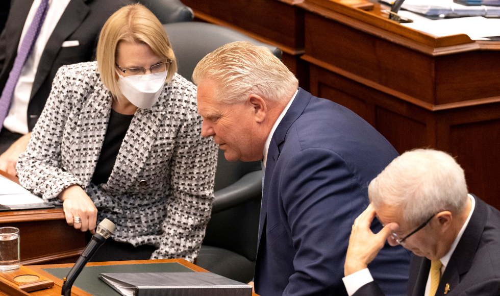 Sylvia Jones wearing a mask leans toward Doug Ford [unmasked] to talk in the legislature.