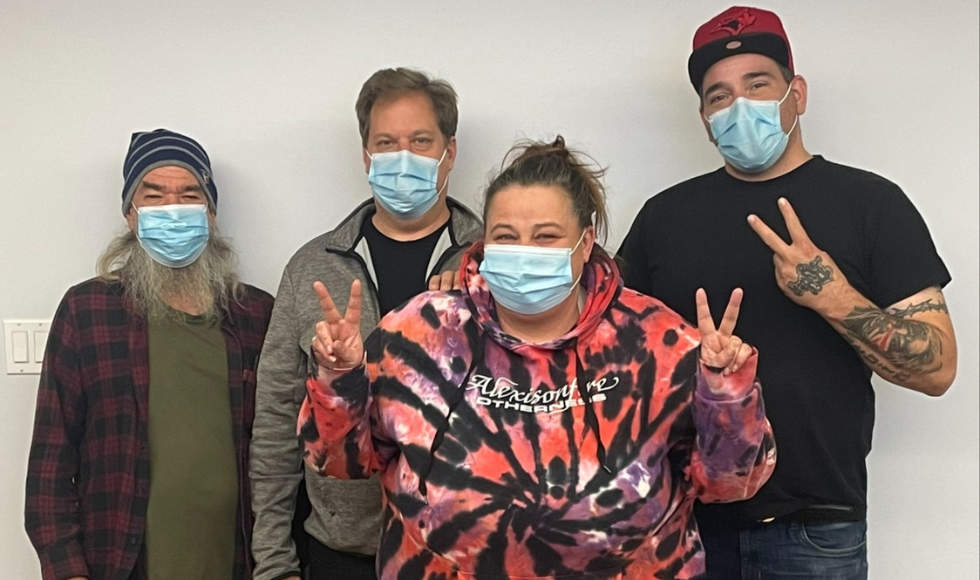 Four masked people photographed indoors. 2 are making a V sign with their fingers.