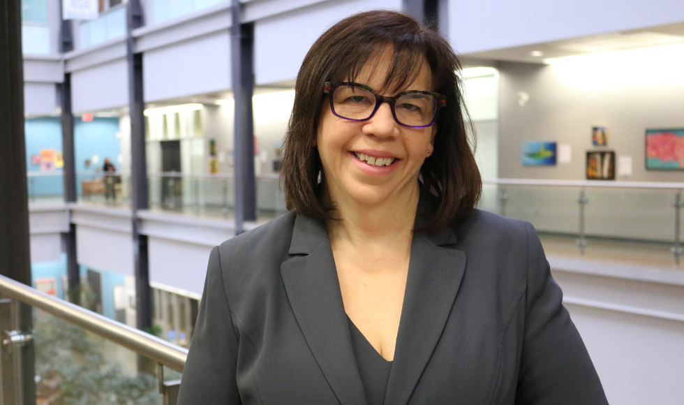 Lauren Griffith standing indoors smiling at the camera. She is wearing black-rimmed glasses and a grey blazer.