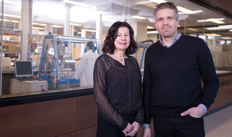 Debi and Hendrick Poinar posing for a photo. Behind them there is a person in a white lab coat working in a laboratory.