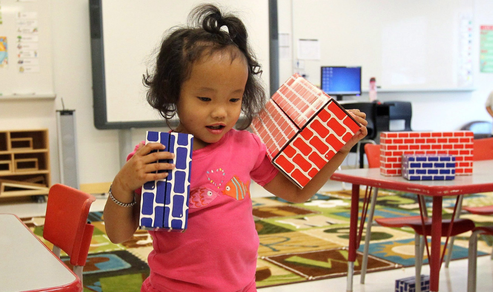 A small child holding toy bricks in a brightly coloured classroom or daycare room.