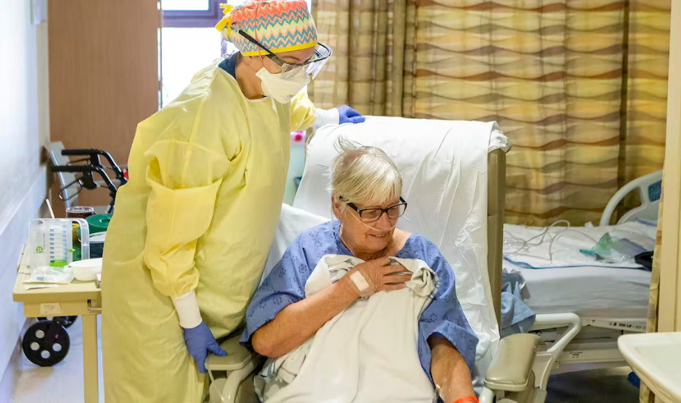 A healthcare worker wearing personal protective equipment standing over a woman seated in a chair inside a hospital room