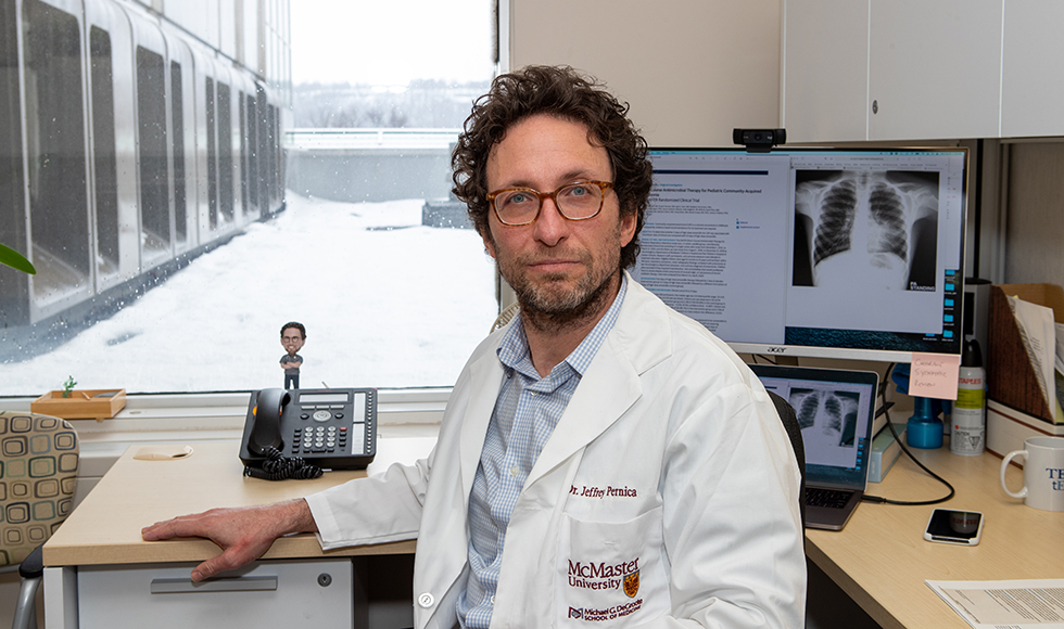 Jeffrey Pernica wearing a white lab coat seated with his back to a desk. On the desk there is a computer that has an X-ray image of a lung open on it.