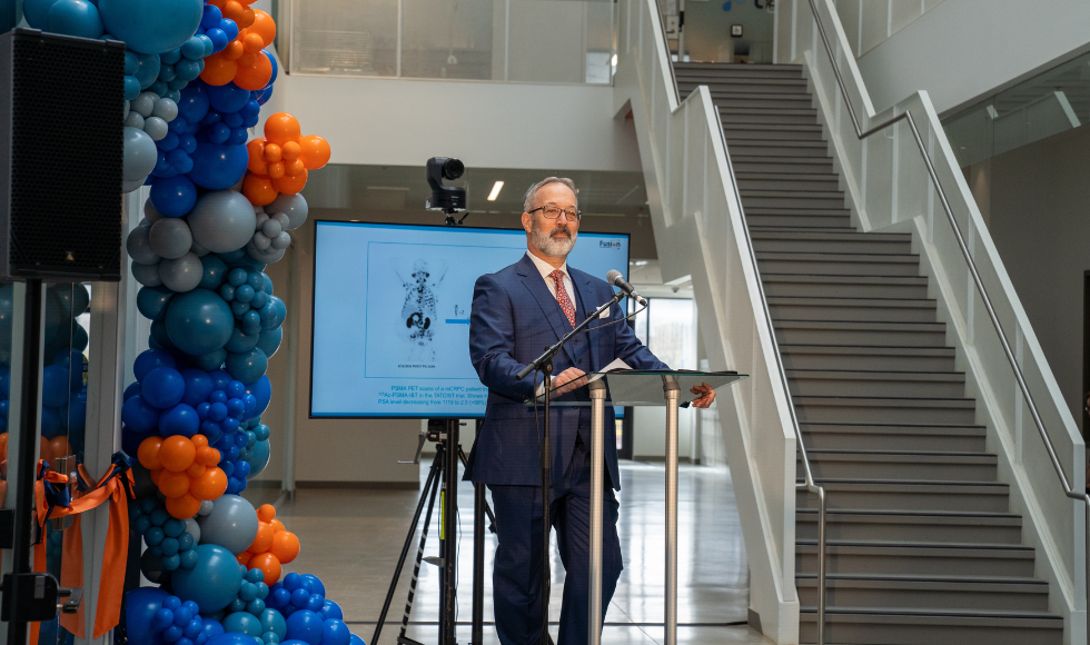 John Valliant speaking at a podium on a stage decorated with blue and orange balloons