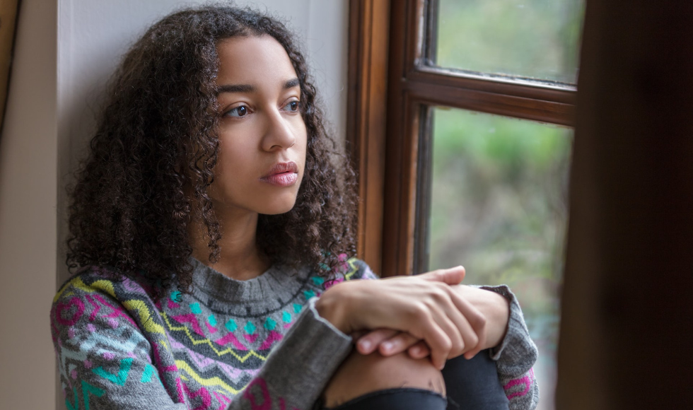 A young girl seated by a window with her hands resting on her knees while looking off in the distance