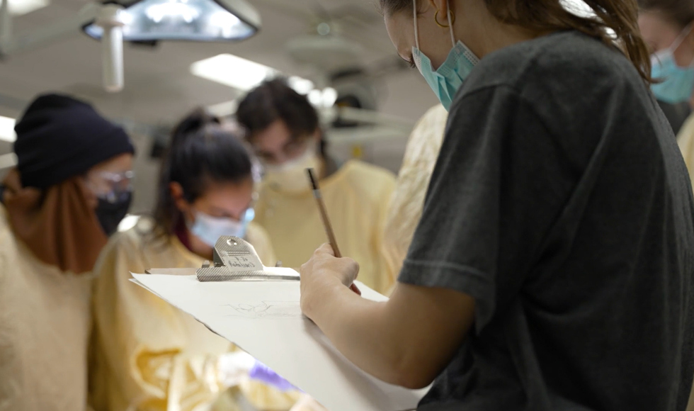 A student drawing on a piece of paper attached to a clipboard. Out of focus in the background there are several students in personal protective gear looking down at something out of sight.