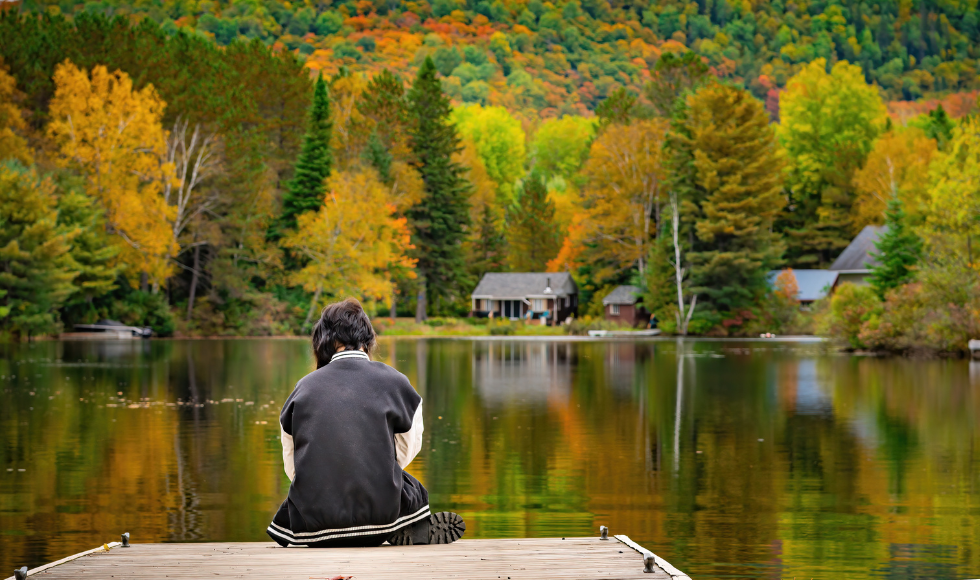 A person seen from behind, sitting at the edge of a dock. the lake or pond is surrounded by trees in fall colours.