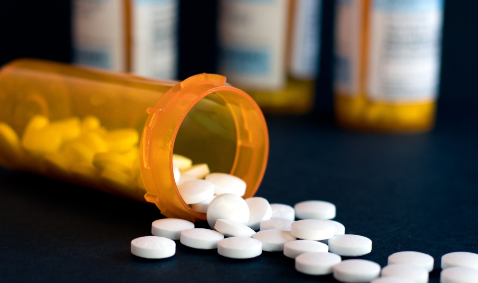 Closeup of a prescription bottle on its side with white pills spilling out on to a tabletop.