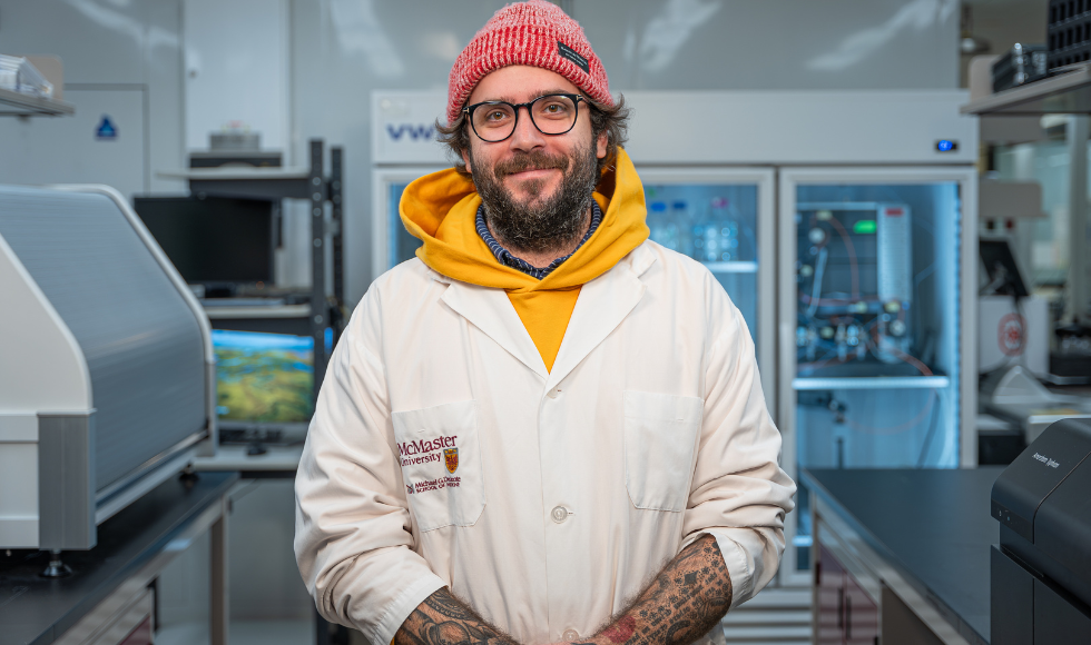 John Stokes, a bearded tattoed man in a hoodie and a lab coat, with glasses and a toque, smiling in his lab.