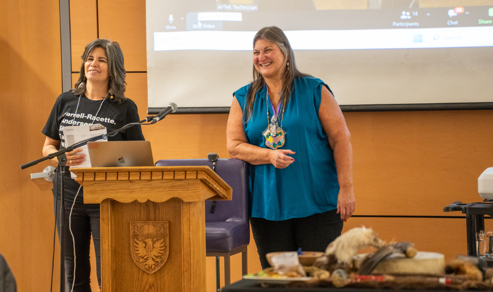 Smiling Savage Bear and Bonnie Freeman standing at a podium in front of a screen with a presentation on it.