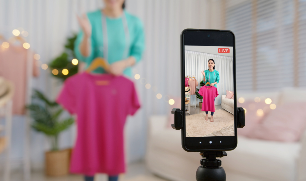 A woman holding up a t-shirt. In the foreground, there is a cellphone showing the woman being live streamed.