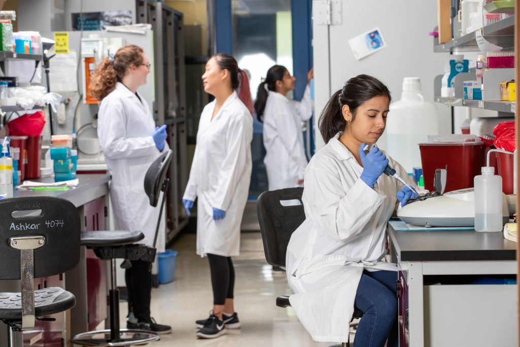 Image of people in a lab, some in the background are talking with one another, and the main person int he image is injecting something into a lab machine.