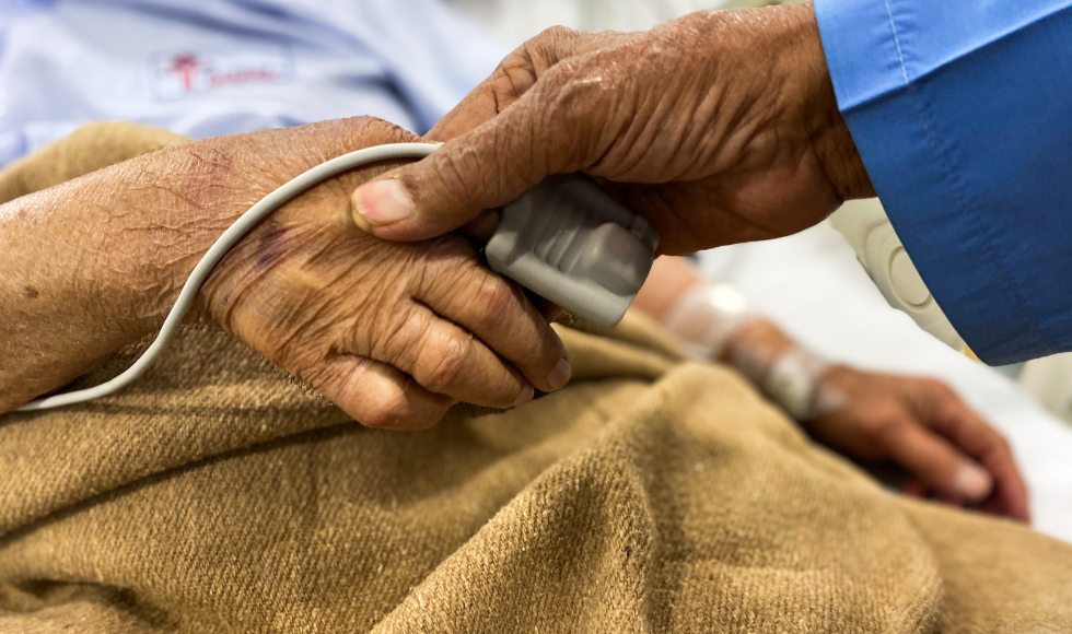 An older couple holds hands. One of them has a heart rate monitor on their finger with a blanket draped over their lap while they sit in a bed.