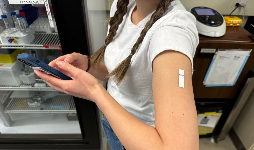 the arms of a person wearing a white short-sleeved Tshirt with a small rectangular patch — smaller than a bandaid — on their arm, holding a smartphone in their hands.