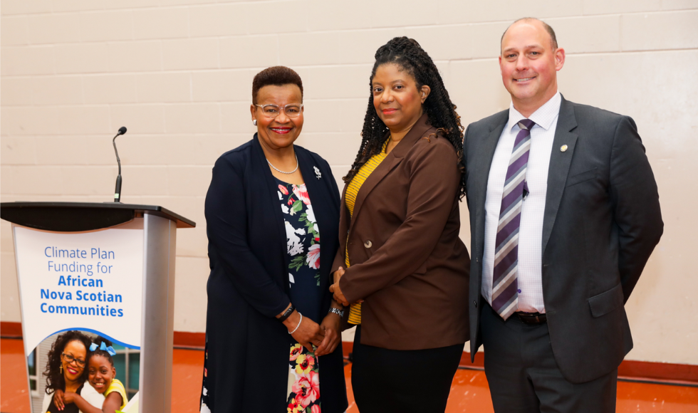 Three people posing for a photo beside a podium. ON the podium there is a photo of a woman and young girl and text that reads, ‘Climate Plan Funding for African Nova Scotian Communities.’