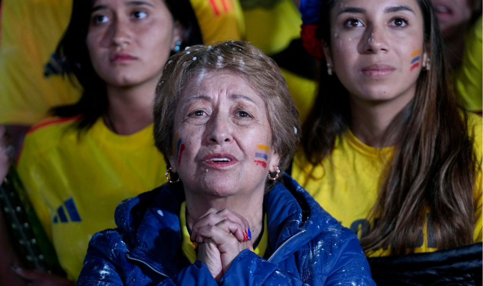 An elderly woman in a blue jacket sits with her hands clasped in front of her