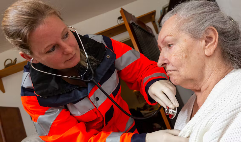 A paramedic wearing a fluorescent jacket holding a stethoscope on the chest of an elderly patient.