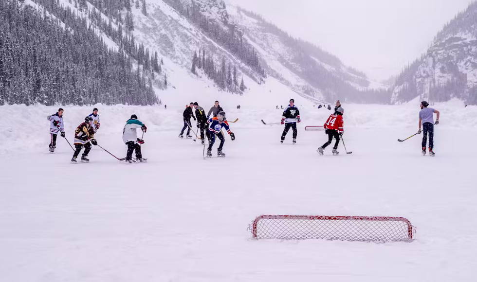 A group of people, most of them in hockey jerseys, playing a hockey on a frozen lake. There are snowy mountains in the background.