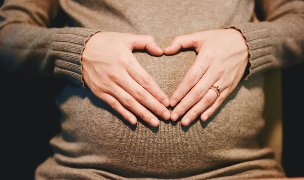 A person wearing a brown top is holding their hands in the shape of a heart on top of their pregnant belly.