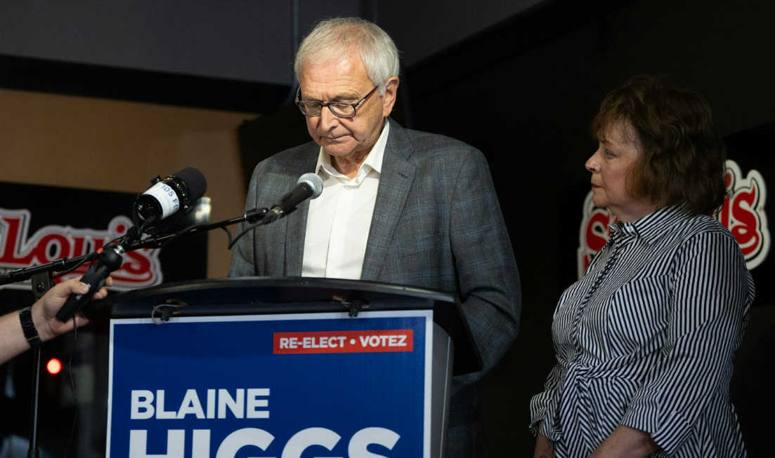 A grey-haired man in glasses looks down as he stands behind a podium that reads Re-elect Blaine Higgs. A dark-haired woman in a striped dress stands beside him.