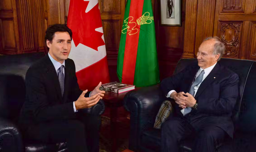 Justin Trudeau and Aga Khan seated on two separate leather couches in a room with ornate wood detailing on the walls. There are two flags behind them.