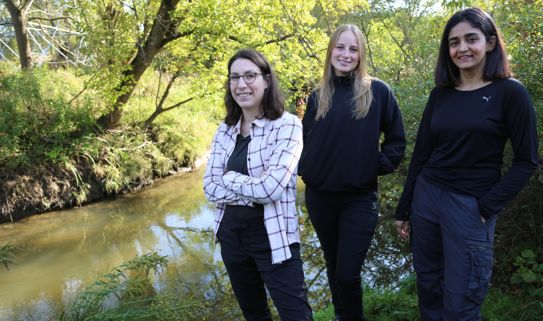 Three smiling people stand an the shore of a tree-lined creek on a summer day.