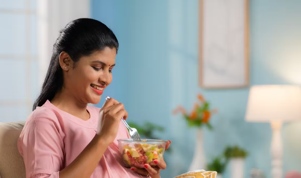 A woman in a pink shirt seated using a fork to eat from a glass bowl of fruit