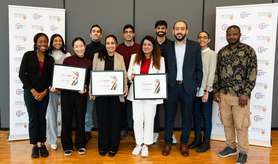 A group of smiling students pose for a picture indoors with McMaster-branded banners on either side of them. Three are holding award certificates.