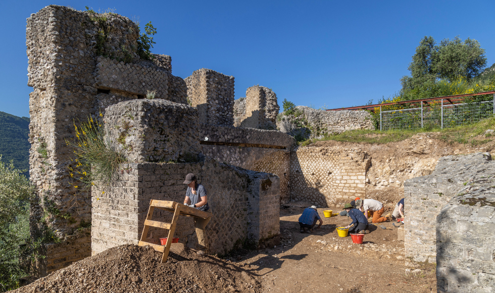 People are working — some standing, some sitting on the ground —  at an archeological dig in the ruins of an old building on a sunny summer day.