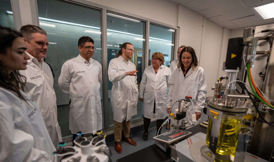 A group of people in lab coats look at lab equipment, standing against the backdrop of a wall of windows.