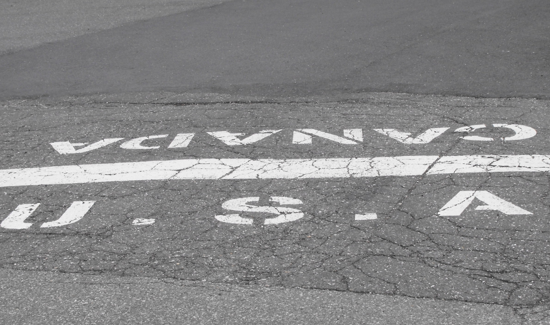 A line of white paint on a roadway separates the U.S. side of the border from the Canadian side. The letters U.S.A. are stencilled on one side of it, 