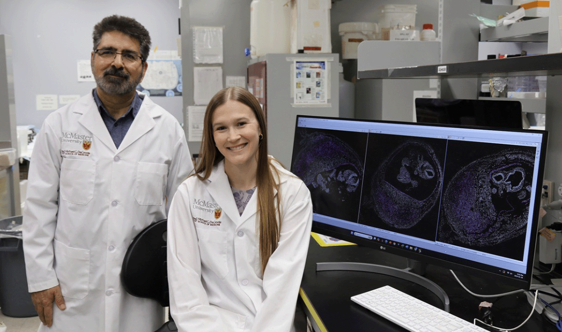 Two people in lab coats inside a lab, beside a monitor showing scans.