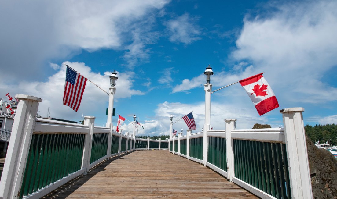 A footbridge stretches ahead with Canadian flags on the rails on one side and U.S. flags on the other.