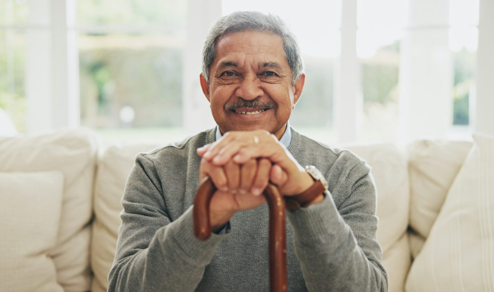 A man sitting on a couch smiles, with his hands resting on a cane in front of his chest. Bright windows are behind him.
