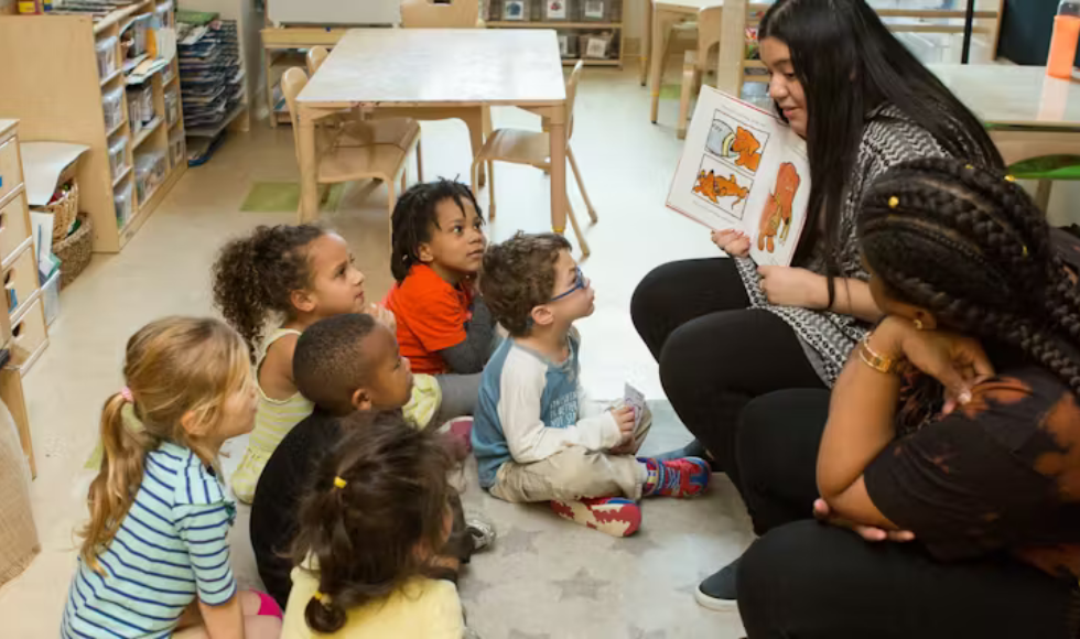 In a classroom, an adult reads a picture book to several children sitting on a mat.